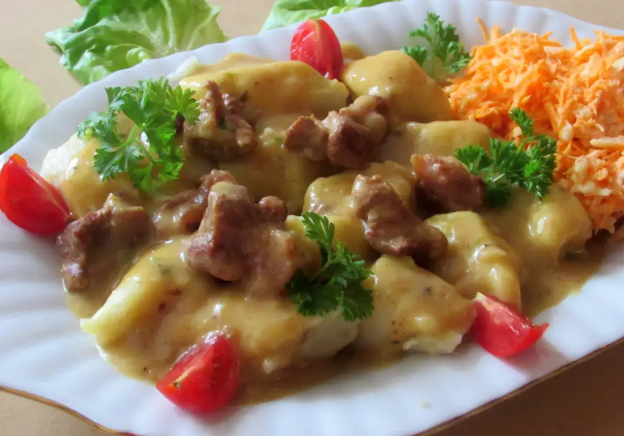 Pyszne kopytka na obiad z gulaszem, polane sosem, udekorowane pomidorkami i natką pietruszki. Obok surówka z marchewki.