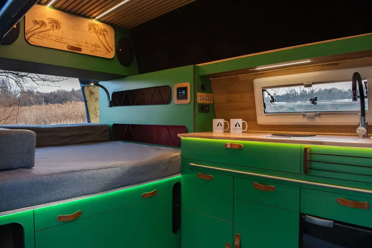 Wnętrze kampera: zielona zabudowa kuchenna, wygodne łóżko, panel sterowania i okno z widokiem na naturę.