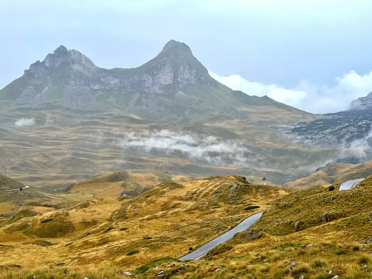 Majestatyczne czarnog&oacute;ra g&oacute;ry w chmurach, z krętą drogą wijącą się przez złote wzg&oacute;rza.