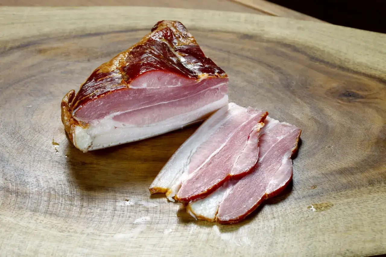 Wędzony boczek pokrojony w plastry na drewnianej desce. Zastanawiasz się, ile czasu parzyć boczek po wędzeniu, by był idealny?