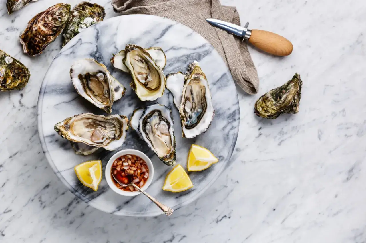 Świeże ostrygi na marmurowej desce, gotowe do jedzenia. Obok sos, cytryna i nóż do ostryg. Idealny sposób, jak jeść ostrygi.