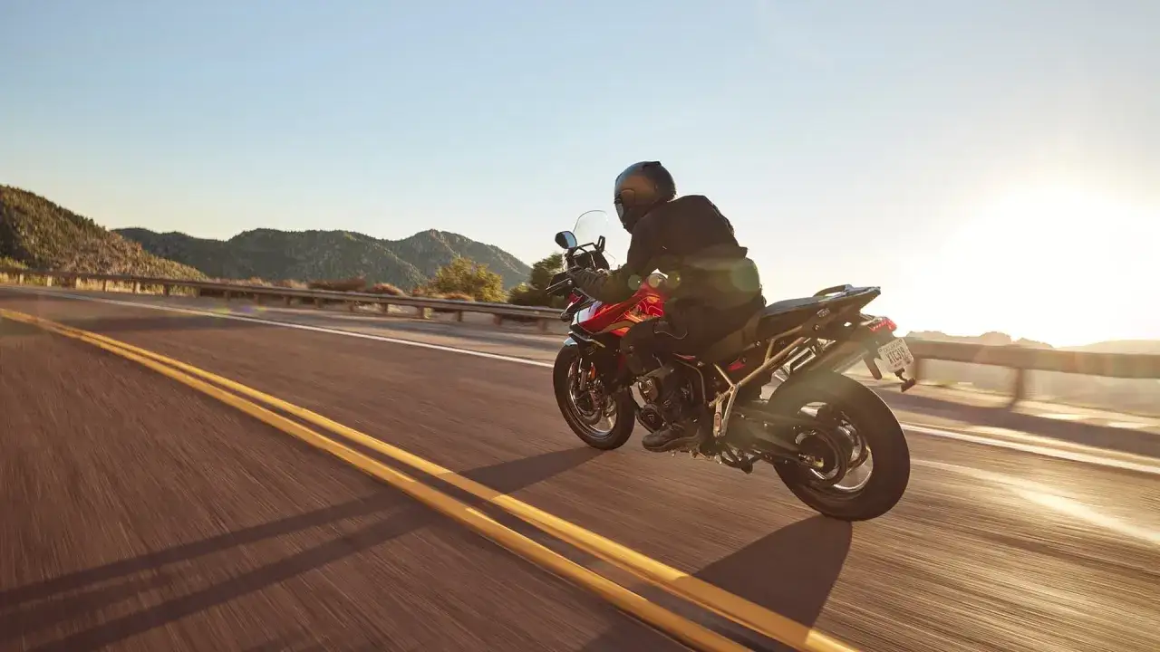 Motocyklista na czerwonym motocyklu Tiger 900 GT Pro przemierza malowniczą g&oacute;rską drogę o zachodzie słońca.