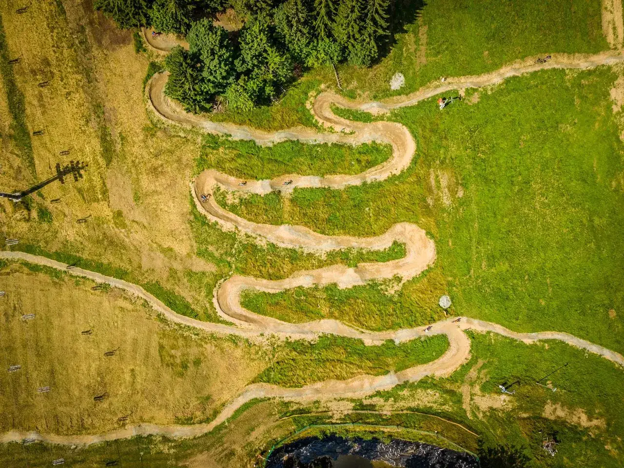 Widok z lotu ptaka na kręte trasy zieleniec bike park, gdzie rowerzyści pokonują zakręty wśród zieleni i suchej trawy.