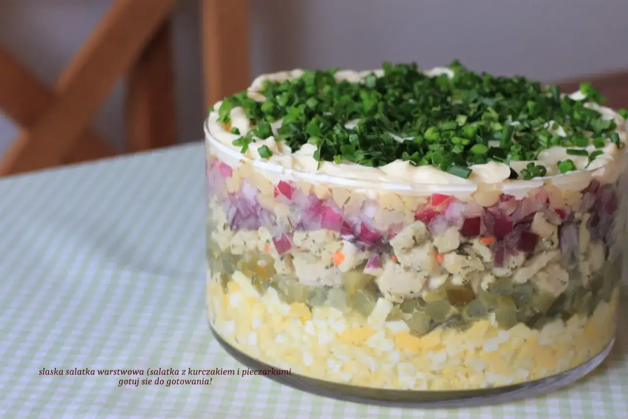 Warstwowa sałatka leśna polana z kurczakiem i pieczarkami, ozdobiona majonezem i szczypiorkiem.