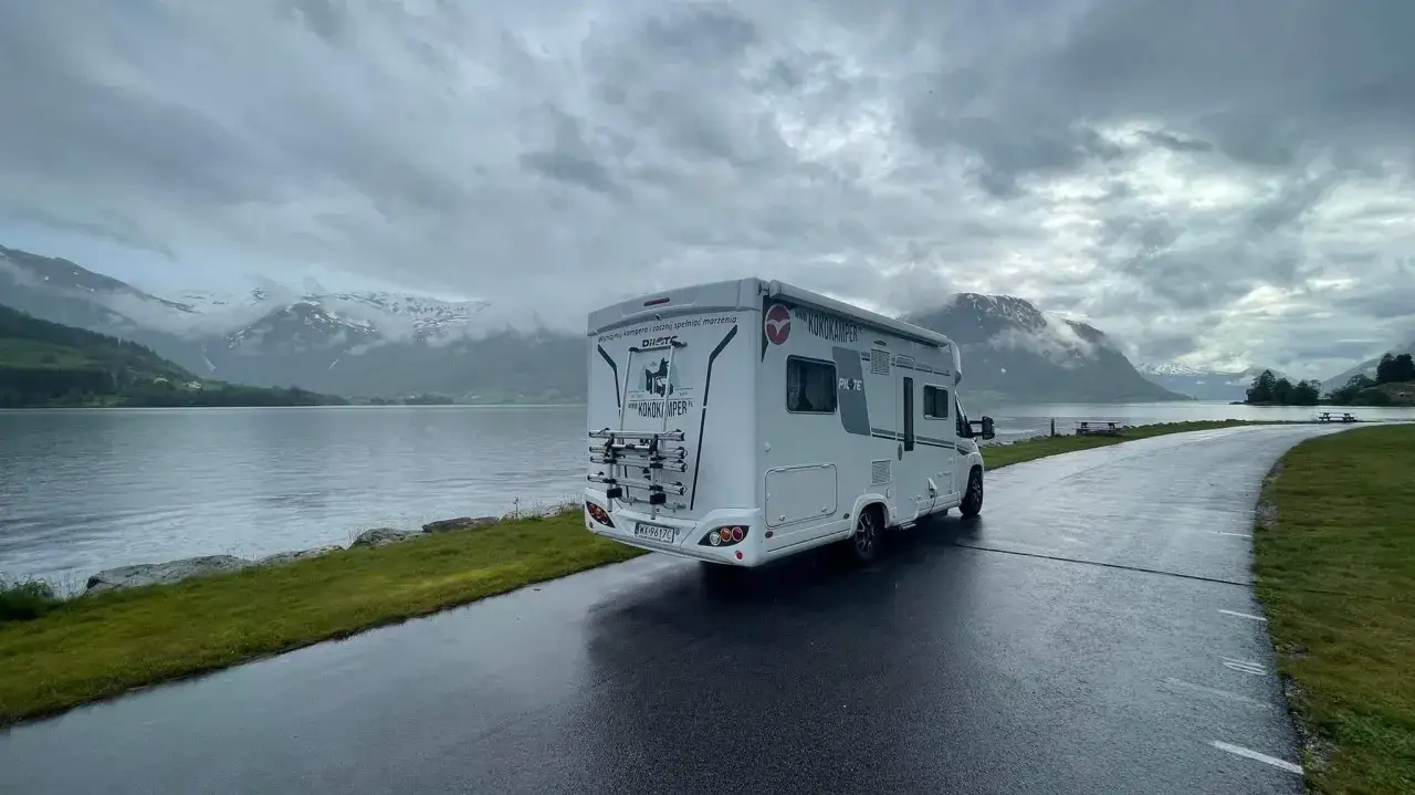 Biały kamper stoi nad jeziorem, w tle góry i pochmurne niebo. Idealne miejsce na podróż kamperem.
