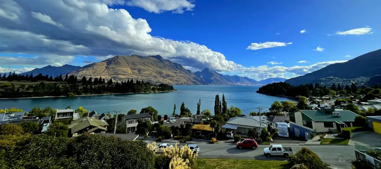 Widok na jezioro i g&oacute;ry w Nowej Zelandii. W oddali widać pasmo g&oacute;rskie, a bliżej domy i drzewa. To wspaniałe nowa zelandia atrakcje.