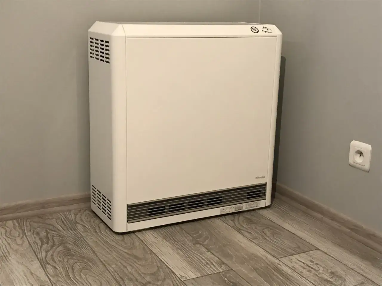 Biały piec akumulacyjny elektryczny stoi w rogu pokoju na drewnianej podłodze.