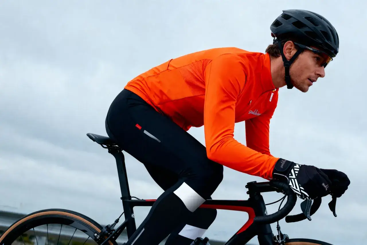 Kolarz w pomarańczowej bluzie i czarnych spodenkach, z kaskiem na głowie, trzyma kierownicę. Dobre **jakie rękawiczki na rower** zapewniają komfort i przyczepność.