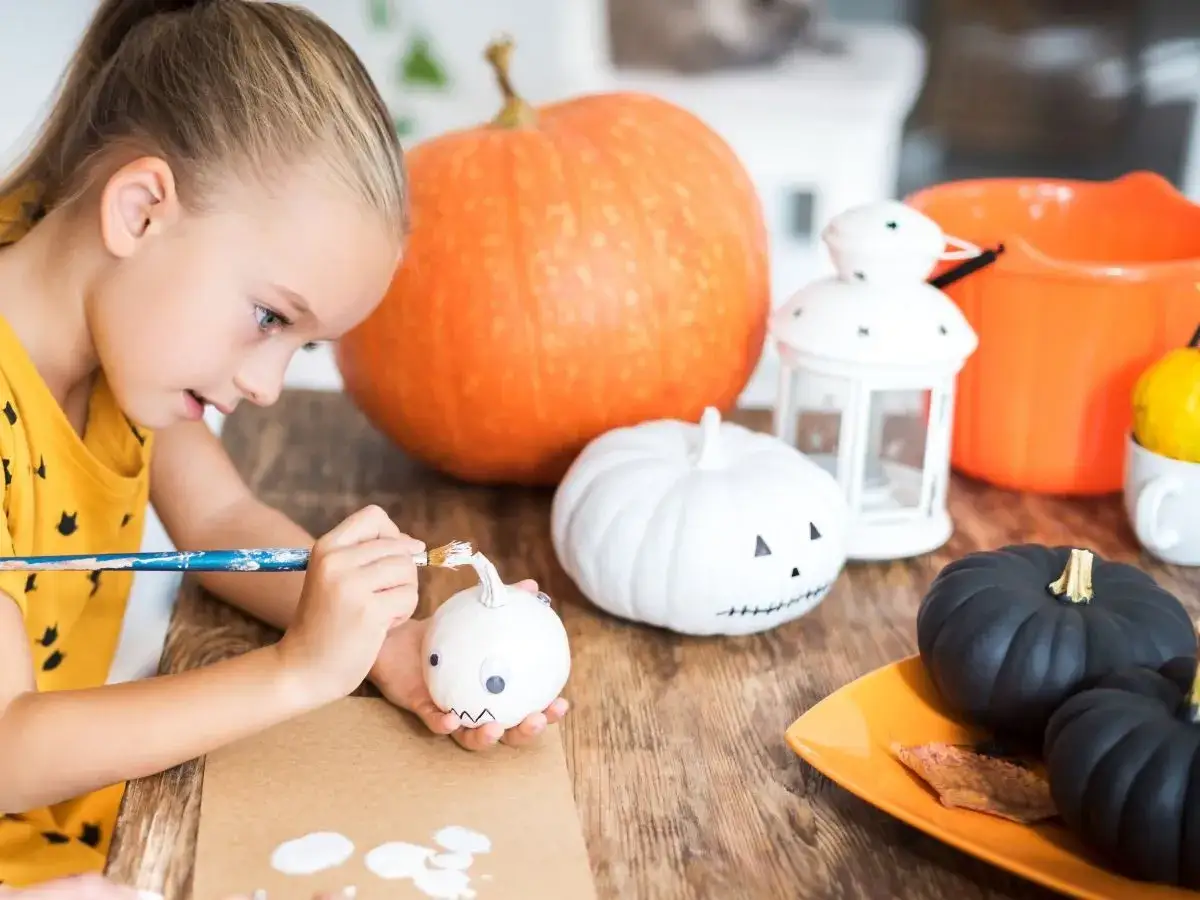 Dziewczynka maluje małą białą dynię, tworząc pomysł na dynię do przedszkola. Obok leżą czarne i pomarańczowe dynie.