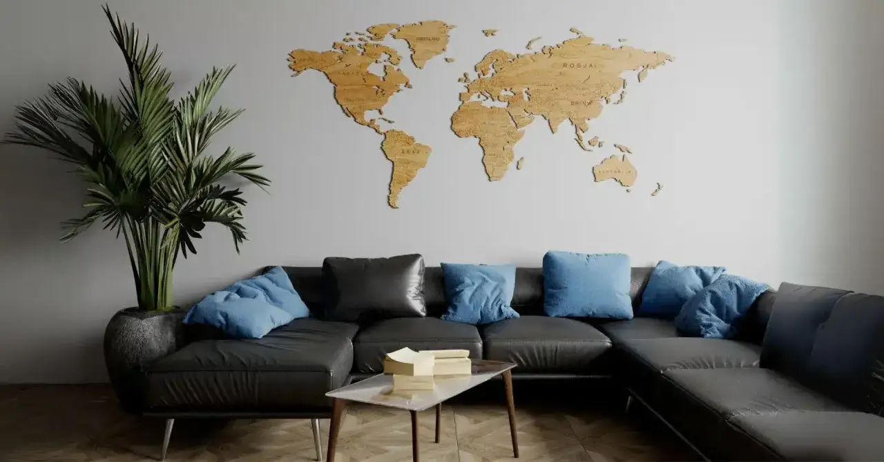 Drewniana mapa świata na ścianie to świetny spos&oacute;b, jak ozdobić ścianę w pokoju. Obok czarna kanapa z niebieskimi poduszkami i duża roślina.