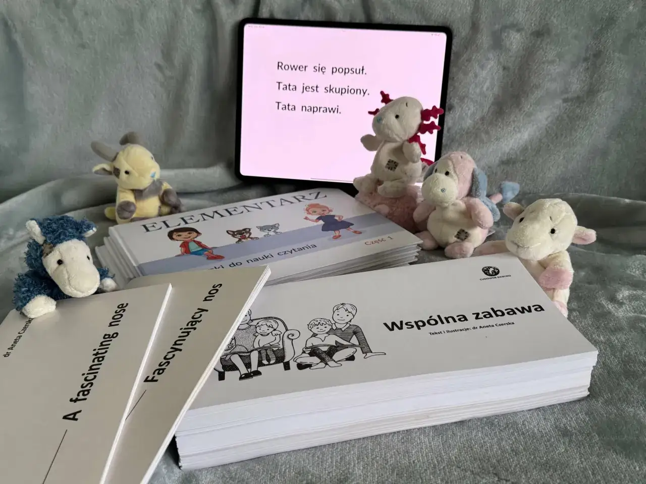 Pluszaki otaczają książki do nauki mówienia i czytania. Na ekranie tabletu wyświetla się tekst, a obok leżą materiały edukacyjne.