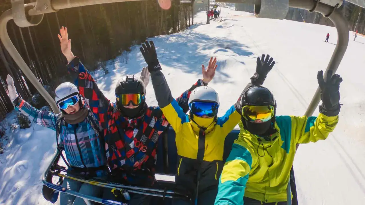 Cztery osoby w kaskach i goglach narciarskich na wyciągu krzesełkowym, machają rękami. W tle widać stok narciarski Budzowski Wierch.