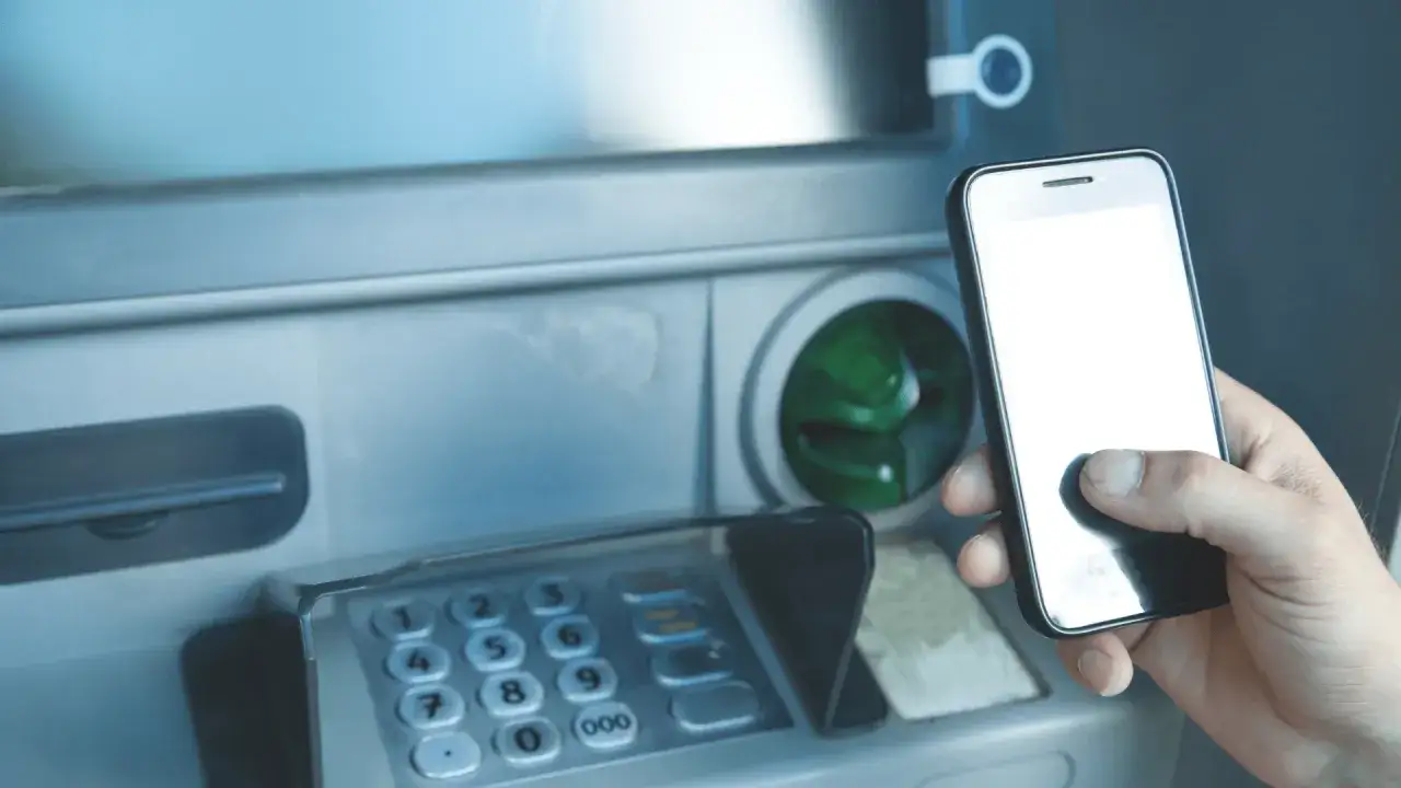 Dłoń trzyma smartfon przy bankomacie, sugerując, czy da się wyplacić pieniadze bez karty.