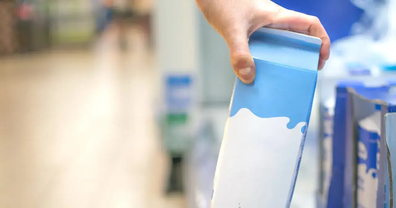 Dłoń trzyma karton po mleku, zastanawiając się, gdzie wyrzucić karton po mleku. W tle widać regały sklepowe.