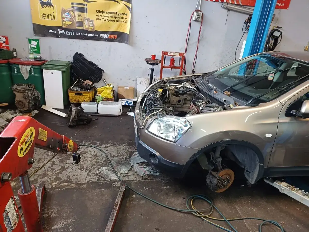 Warsztat samochodowy, gdzie trwa wymiana silnika w samochodzie. Widać rozebrany prz&oacute;d auta, skrzynię bieg&oacute;w i silnik na podnośniku.