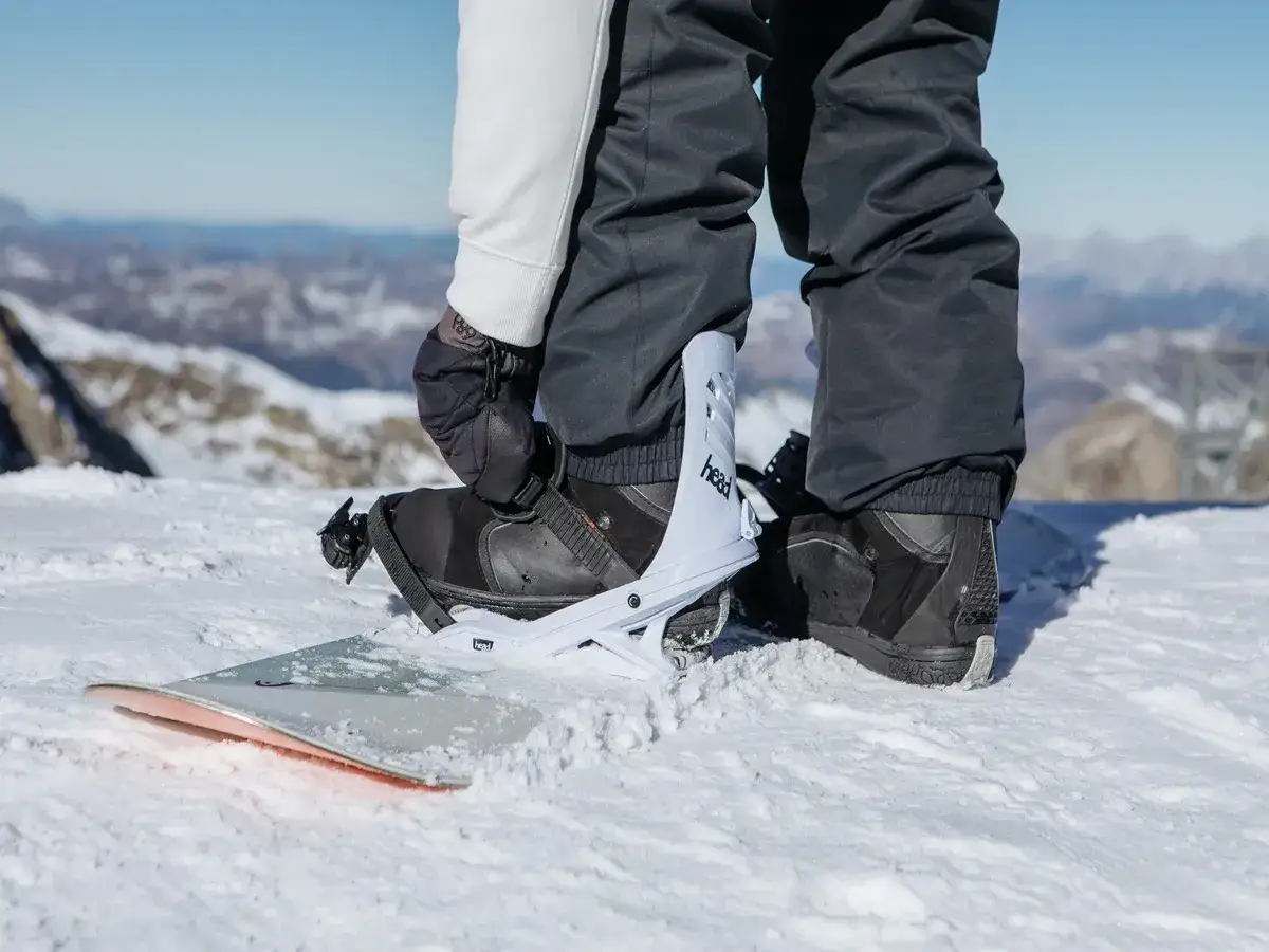Osoba przygotowuje się do jazdy na snowboardzie. Czy buty narciarskie pasują do snowboardu? Nie, to specjalne buty snowboardowe wpięte w wiązania.