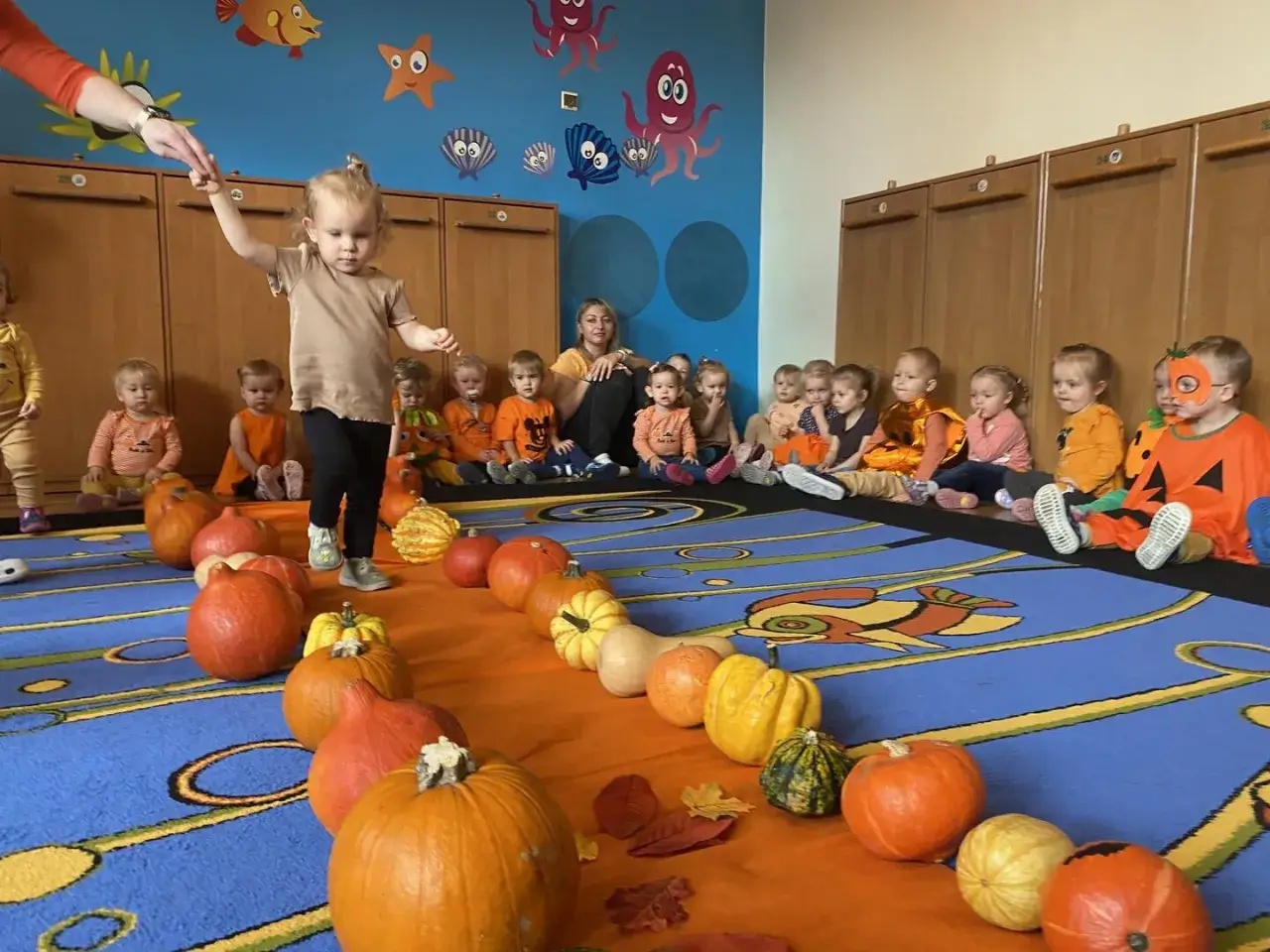 Dzieci w przedszkolu świętują jesień, tworząc pomysł na dynię do przedszkola. Rząd dyni i małych przedszkolaków w strojach.