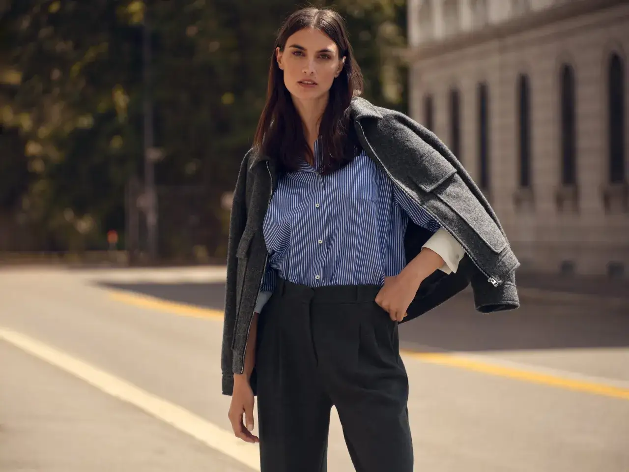 Kobieta w granatowo-białej koszuli w paski i grafitowych spodniach. Szara marynarka narzucona na ramiona. Jaki kolor pasuje do niebieskiego ciemnego? Szarość jest doskonałym wyborem.