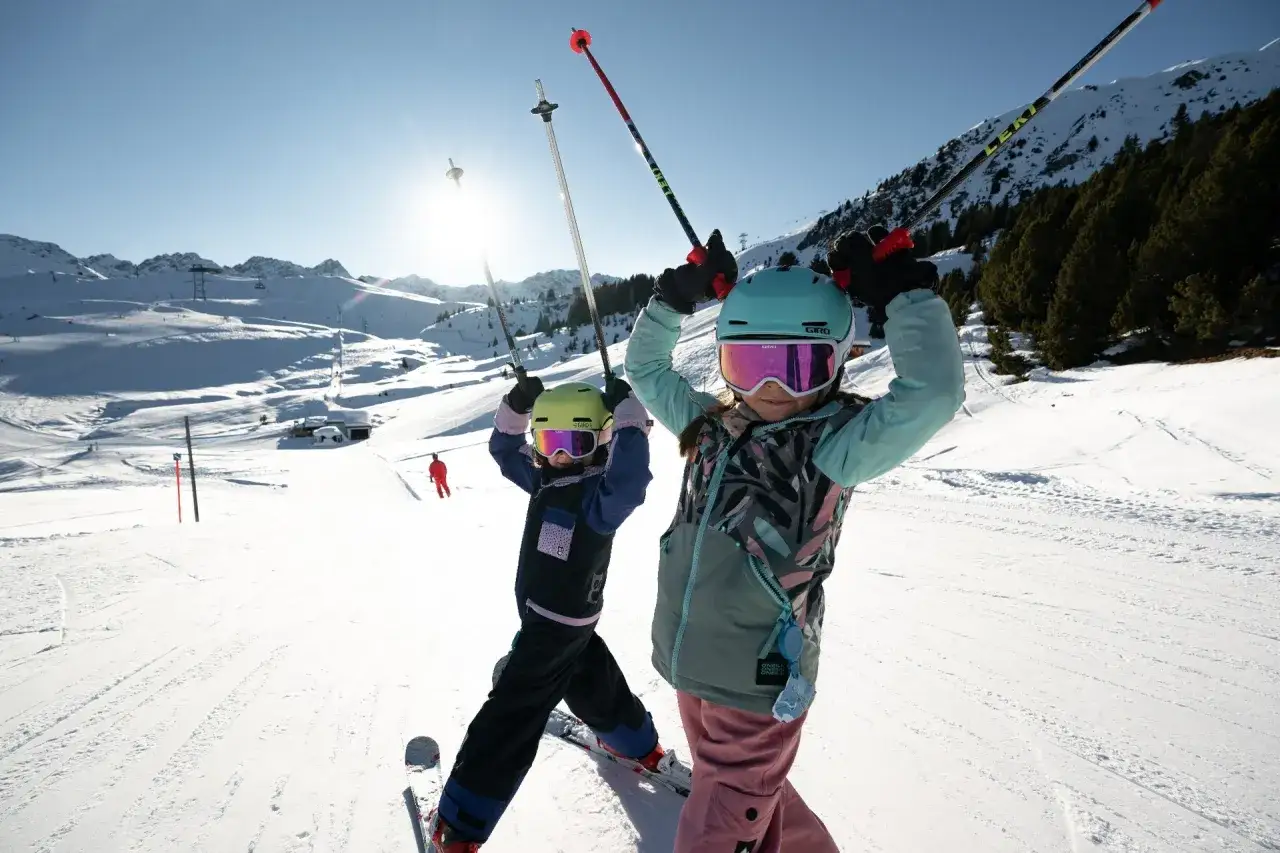 Dwie dziewczynki w kaskach i goglach cieszą się jazdą na nartach. Młodsza, ubrana na ciemno, ma narty dla 3 latka. Słońce świeci na zaśnieżonym stoku.