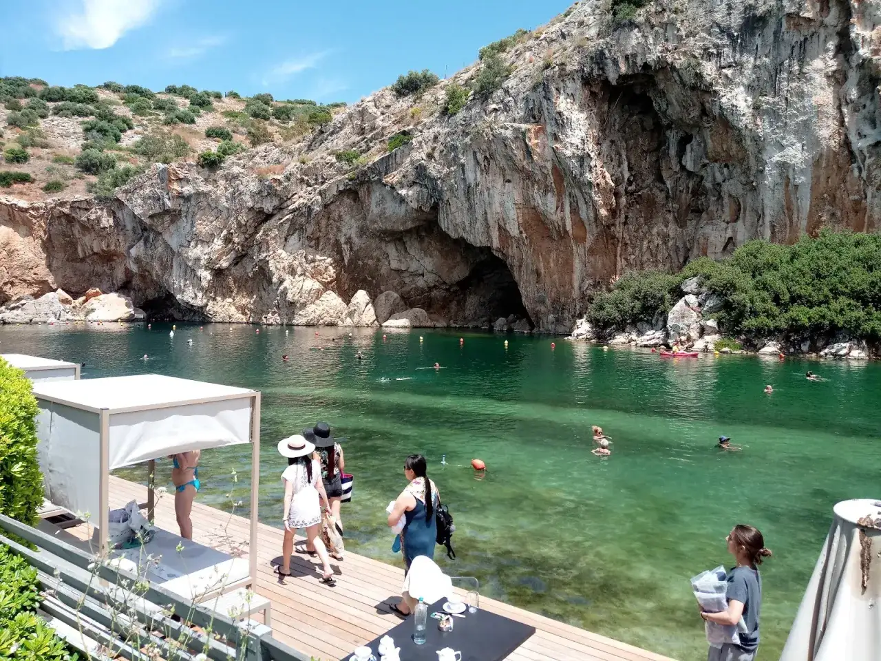 Słoneczny dzień nad jeziorem Vouliagmeni. Ludzie pływają w krystalicznie czystej wodzie, a skalne ściany tworzą malownicze tło.