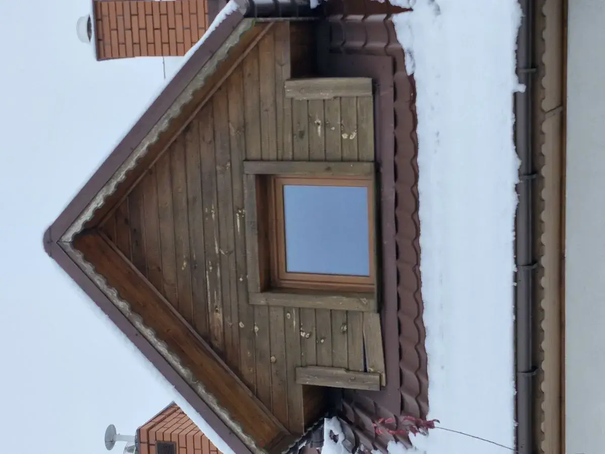 Drewniana ściana szczytowa z oknem, pokryta śniegiem. Widoczna wentylacja w ścianie szczytowej.