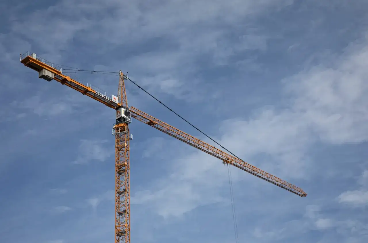 Pomarańczowy żuraw wieżowy na tle błękitnego nieba z chmurami. Operator żurawia wieżowego czuwa nad pracami budowlanymi.