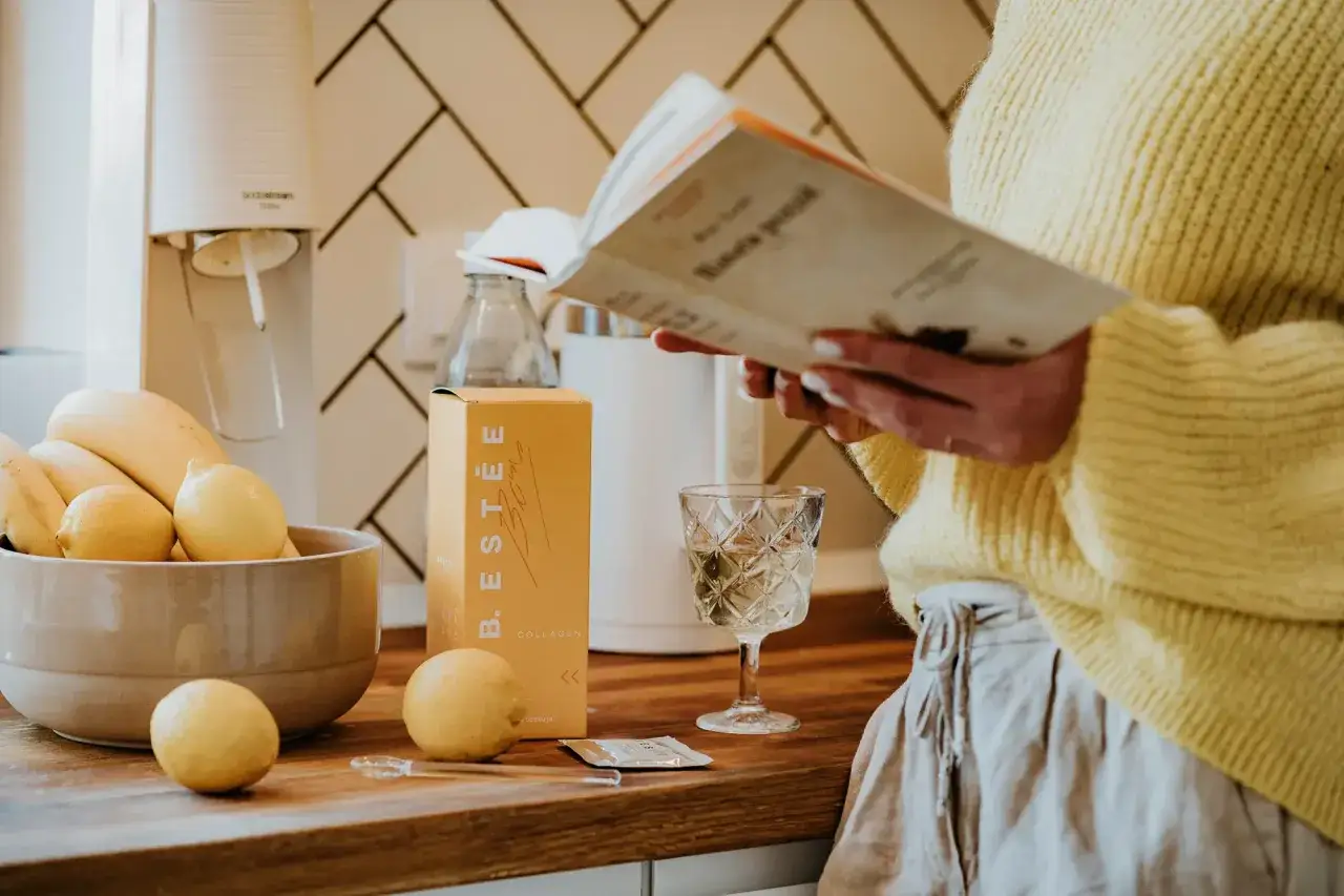 Fotografia produktu: ż&oacute;łte pudełko B.EST&Eacute;E Collagen, cytryny, banany, szklanka wody i czytająca osoba.