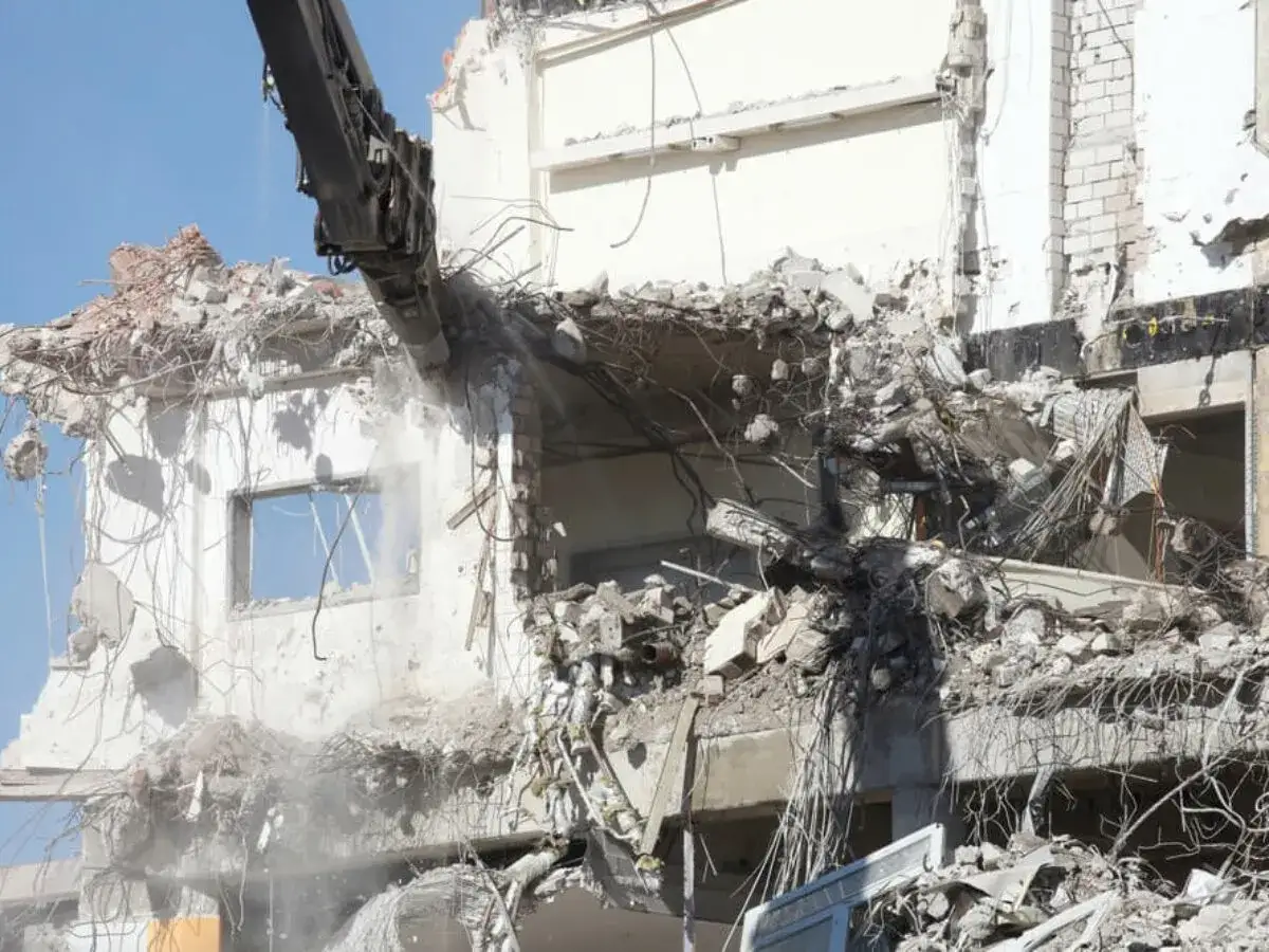 Maszyna budowlana niszczy budynek grożący zawaleniem. W powietrzu unosi się pył, a z gruzów wystają zbrojenia.