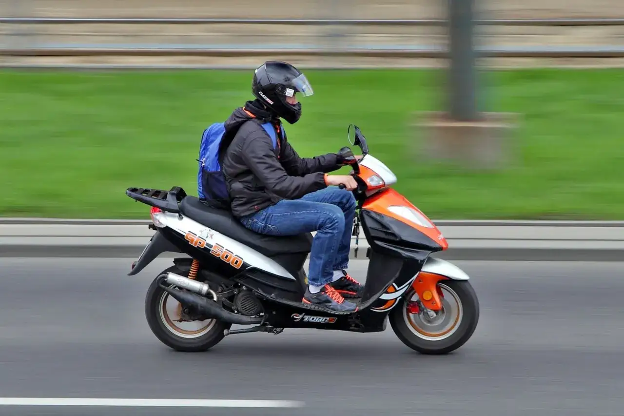 Młody mężczyzna w kasku i z plecakiem jedzie skuterem. Kto może jeździć skuterem? Odpowiedź jest prosta: każdy, kto ma uprawnienia i czuje się pewnie na drodze!