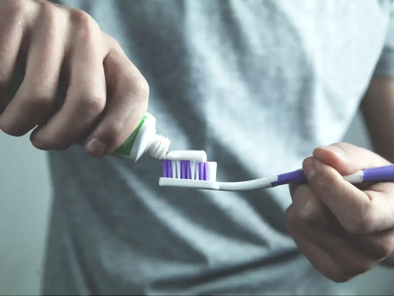 Ręce nakładają pastę do zębów z tubki na szczoteczkę. Pasta zawiera fluor, który chroni zęby.