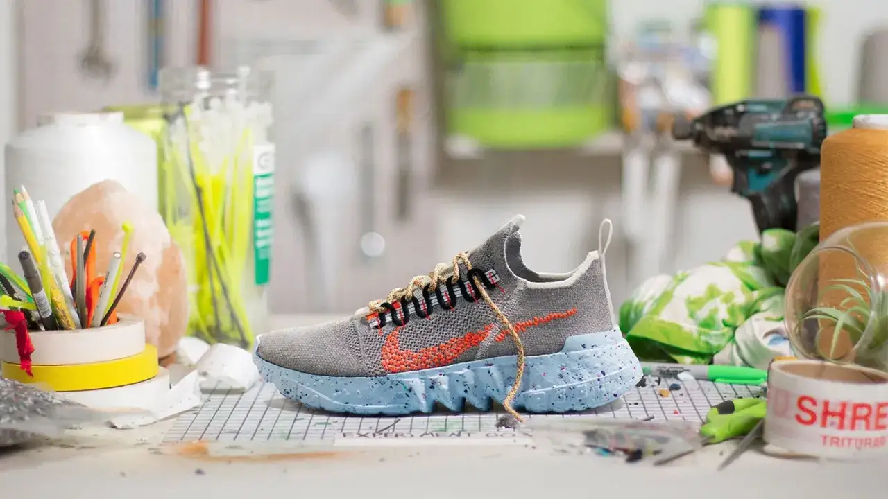 Szare buty Nike z pomarańczowym logo i niebieską podeszwą, wyglądające jak najdziwniejsze buty Nike, leżą na stole z materiałami do tworzenia.