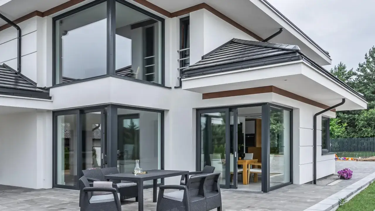 Nowoczesny dom z dużymi oknami, w tym okna otwierane do góry, tarasem i meblami ogrodowymi.