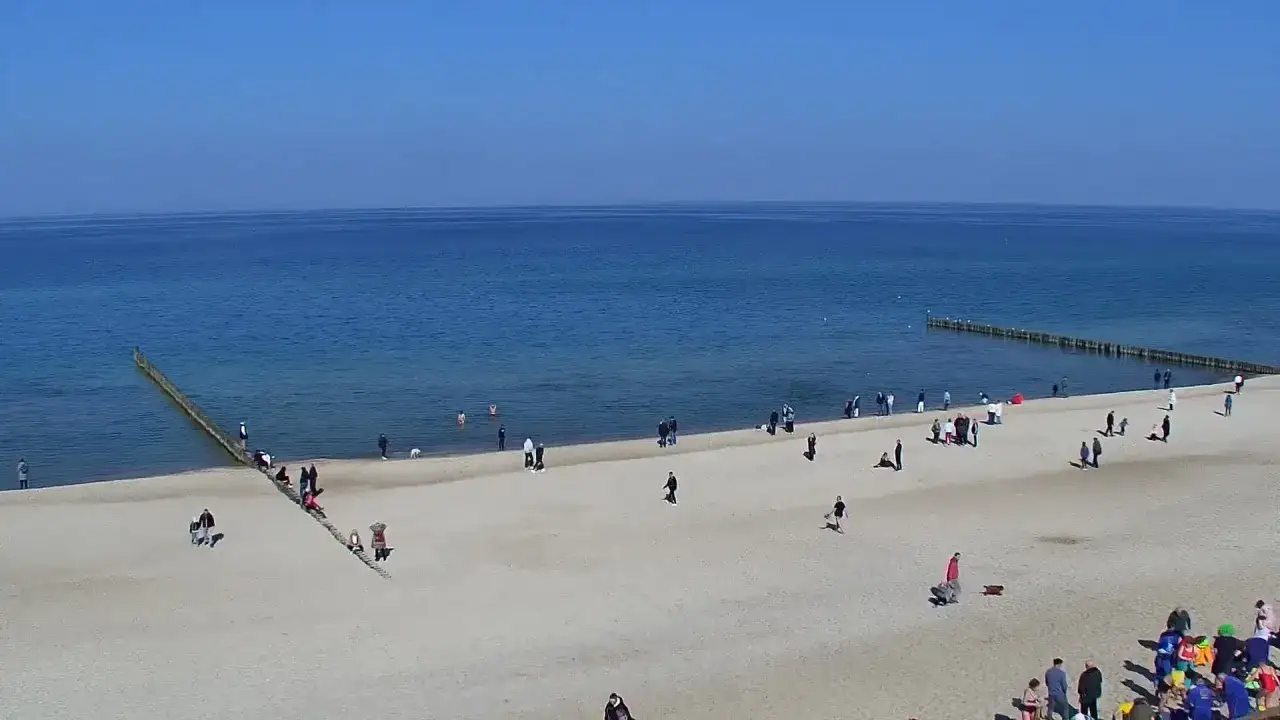 Słoneczny dzień na plaży w Mielnie. Ludzie spacerują, bawią się w wodzie. Ciekawe, ile mieszkańców ma Mielno, że tak wielu ludzi wypoczywa.