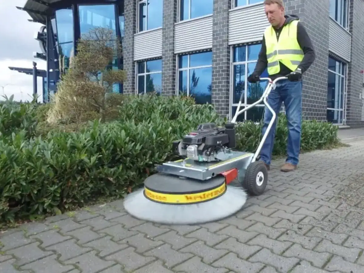 Mężczyzna w kamizelce odblaskowej prowadzi zamiatarkę do kostki brukowej, czyszcząc chodnik przed budynkiem.