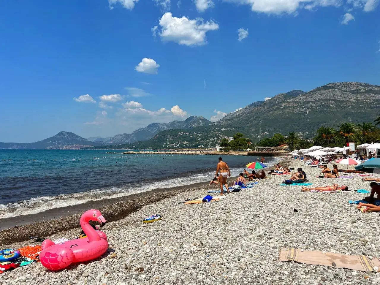 Sutomore atrakcje: plaża z kamykami, morze, g&oacute;ry w tle i mn&oacute;stwo ludzi odpoczywających na ręcznikach. R&oacute;żowy flaming unosi się na wodzie.