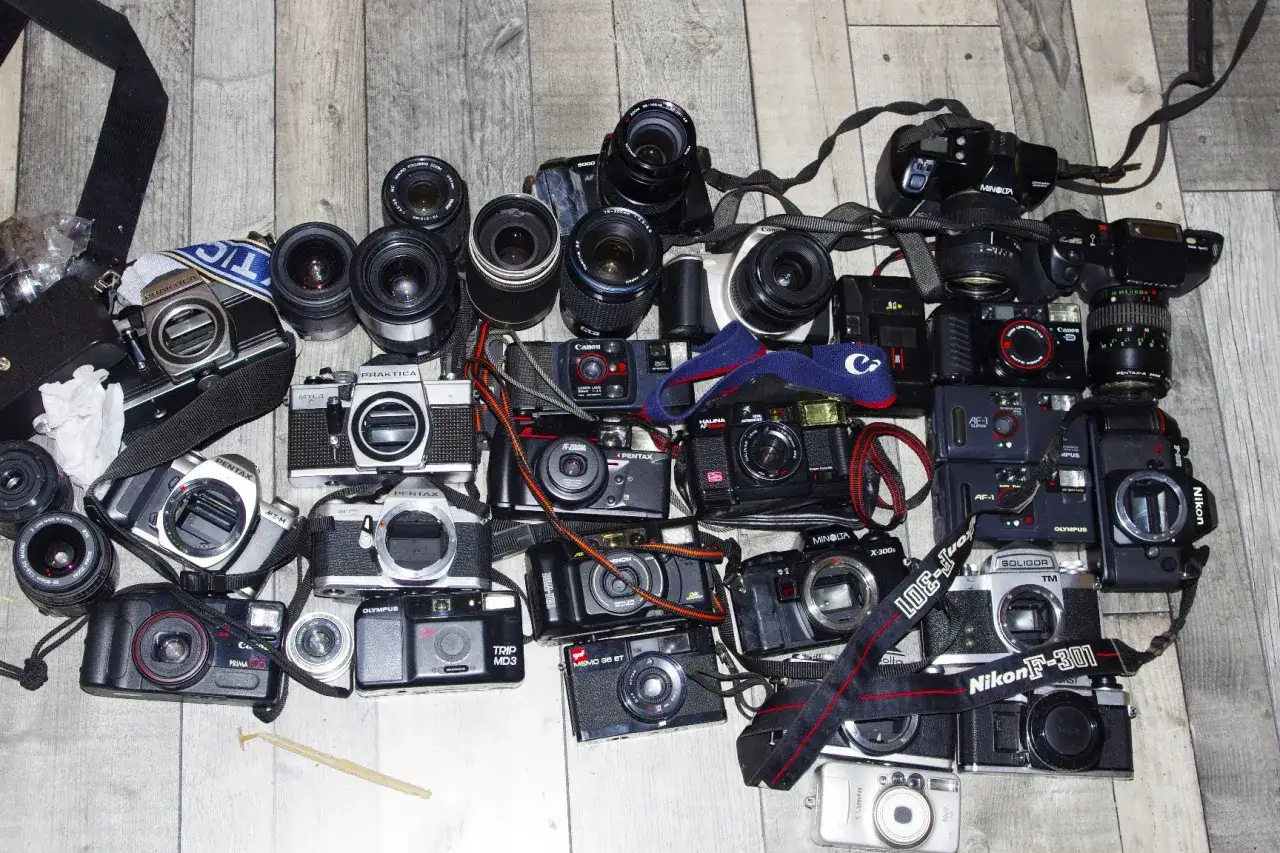 Kolekcja starych aparatów fotograficznych na klisze, w tym modele Pentax, Canon, Nikon, Olympus i Minolta, leżących na drewnianym tle.