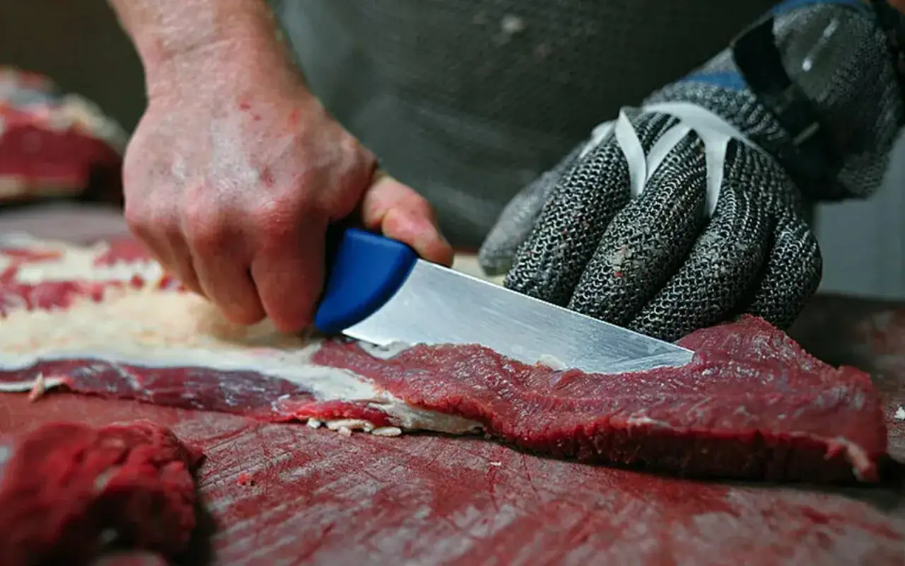Ręce w rękawicy ochronnej i z nożem usuwają błony i ścięgna z surowego mięsa. Proste triki, jak pozbyć się błon i ścięgien z mięsa.
