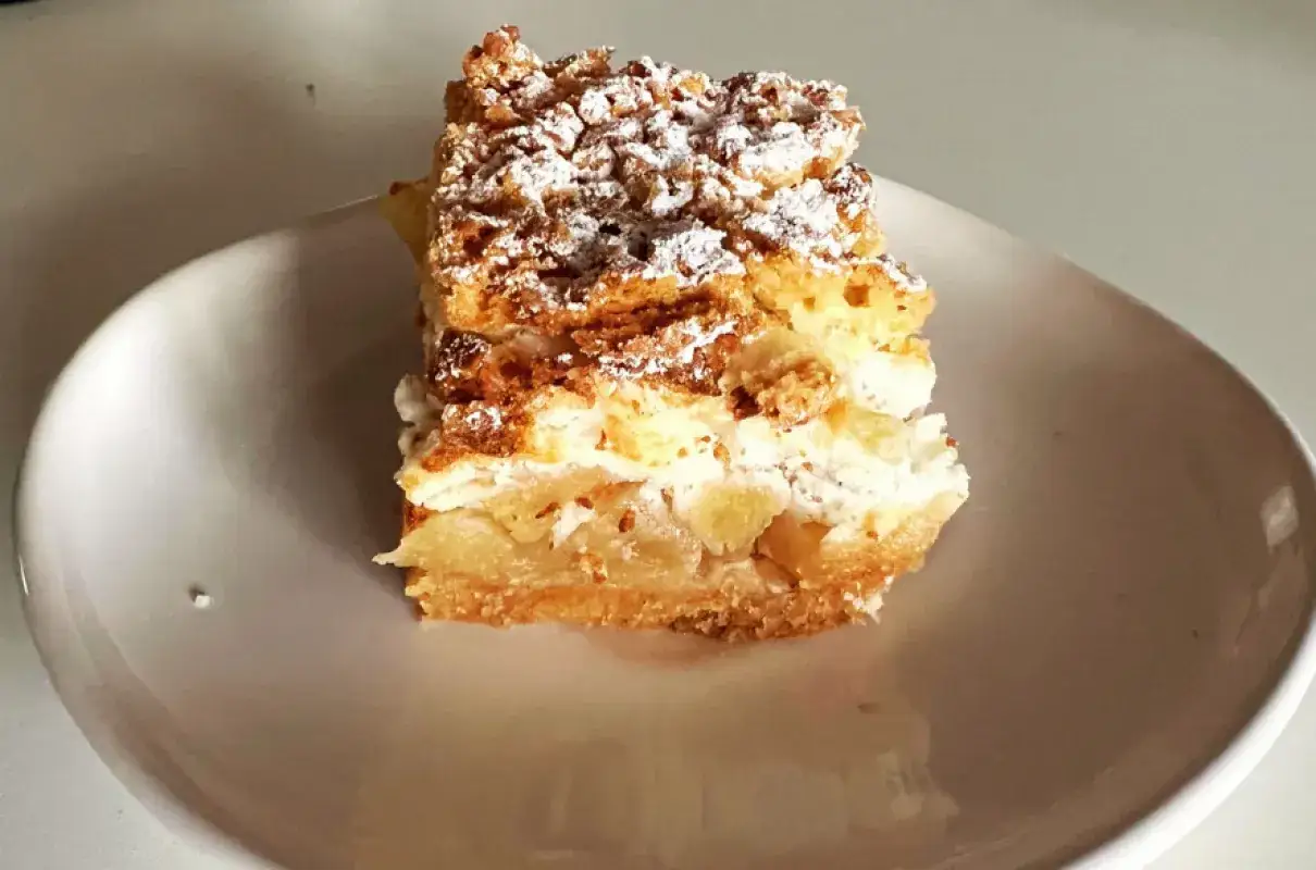 Pyszna szarlotka z bezą i kruszonką, z kawałkami jabłek i cukrem pudrem, na białym talerzu.