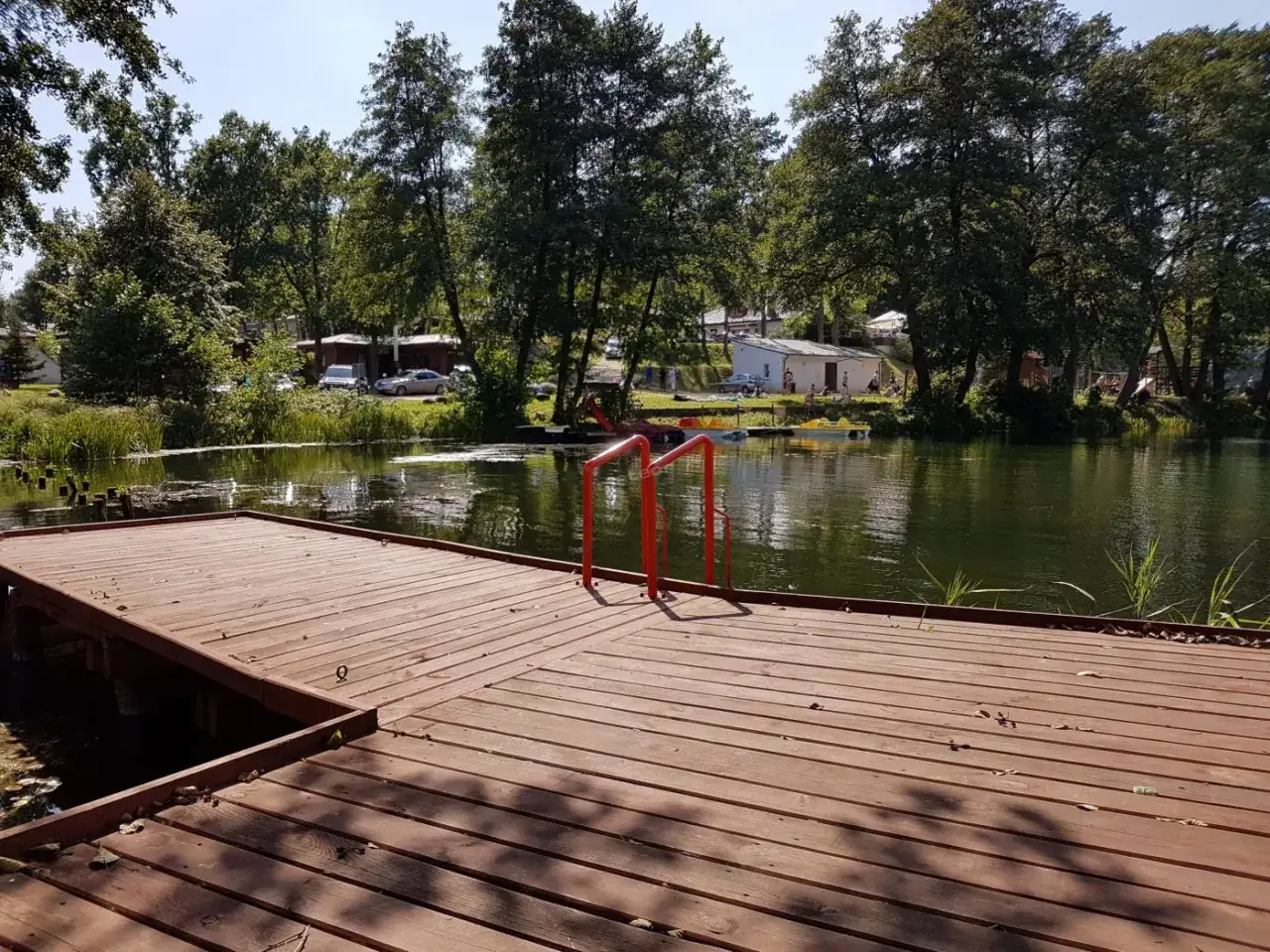 Drewniane molo z czerwonymi poręczami nad jeziorem, w tle drzewa i budynki ośrodka łagów camping.