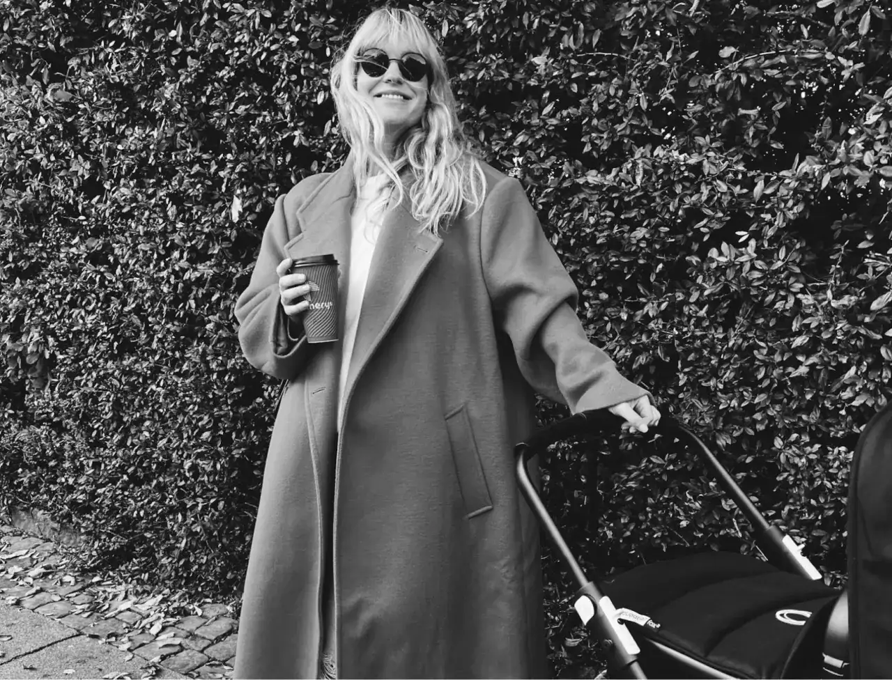 Uśmiechnięta kobieta w płaszczu, z kawą w ręku, prowadzi w&oacute;zek. Stylizacją przypomina bohaterki kampanii skandynawskich firm odzieżowych.