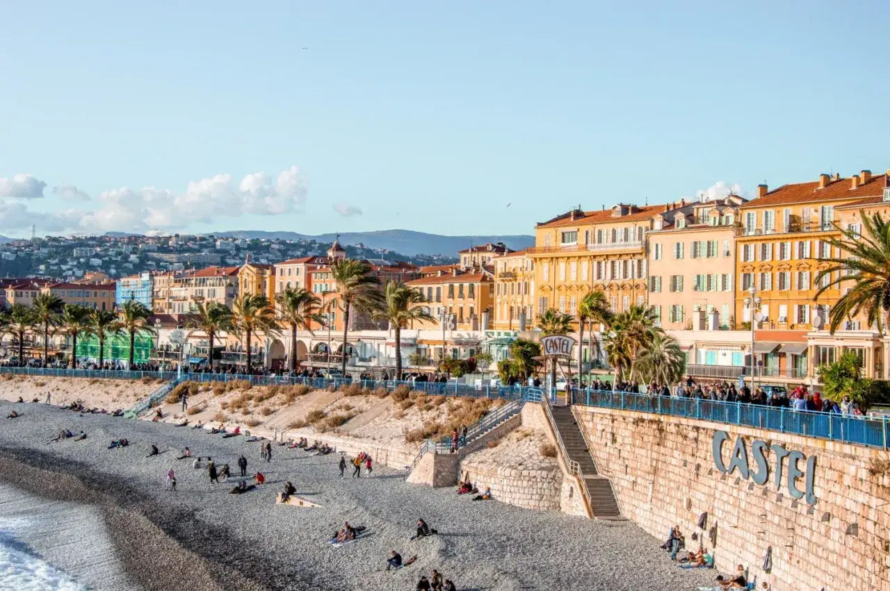 Kamienista plaża w Nicei, z palmami i kolorowymi kamienicami. Idealne miejsce, by zobaczyć w Nicei, jak ludzie odpoczywają nad morzem.