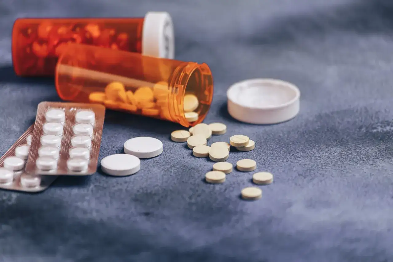 Pigułki, w tym niesteroidowe leki przeciwzapalne, rozsypane z pomarańczowych buteleczek na szarym tle.