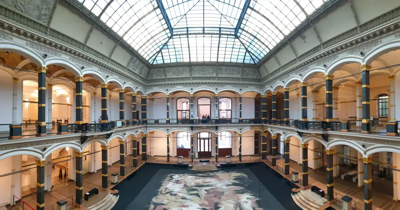 Wnętrze muzeum sztuki nowoczesnej w Berlinie z imponującym, przeszklonym dachem i galerią otaczającą centralną przestrzeń.