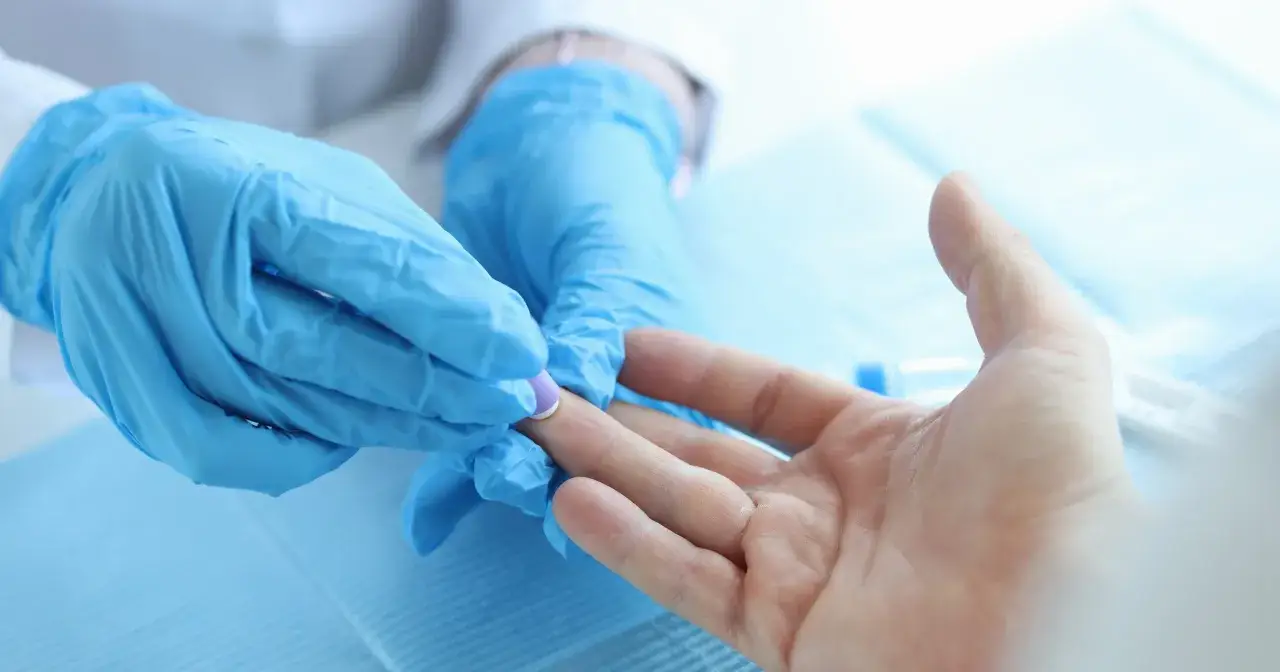 Lekarz w niebieskich rękawiczkach pobiera pr&oacute;bkę krwi z palca pacjenta. Badanie CRP pomaga ocenić stan zapalny.