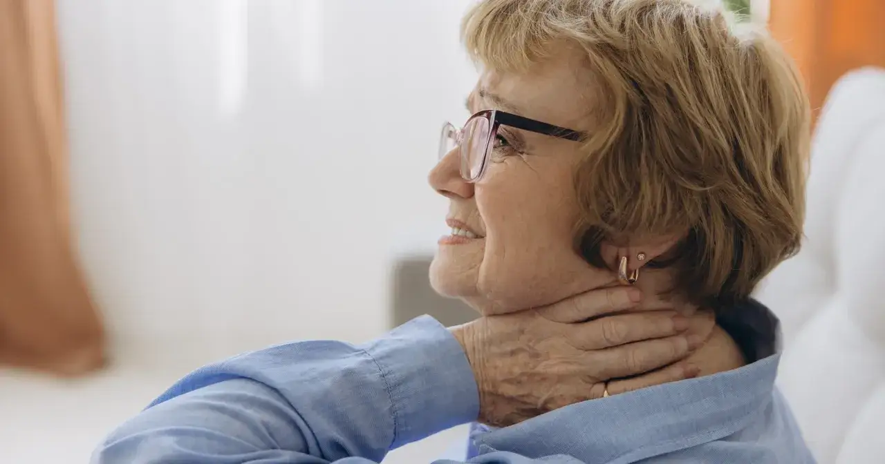 Starsza kobieta w okularach dotyka szyi, co może symbolizować objawy somatyczne, czyli fizyczne odczucia związane ze stresem.