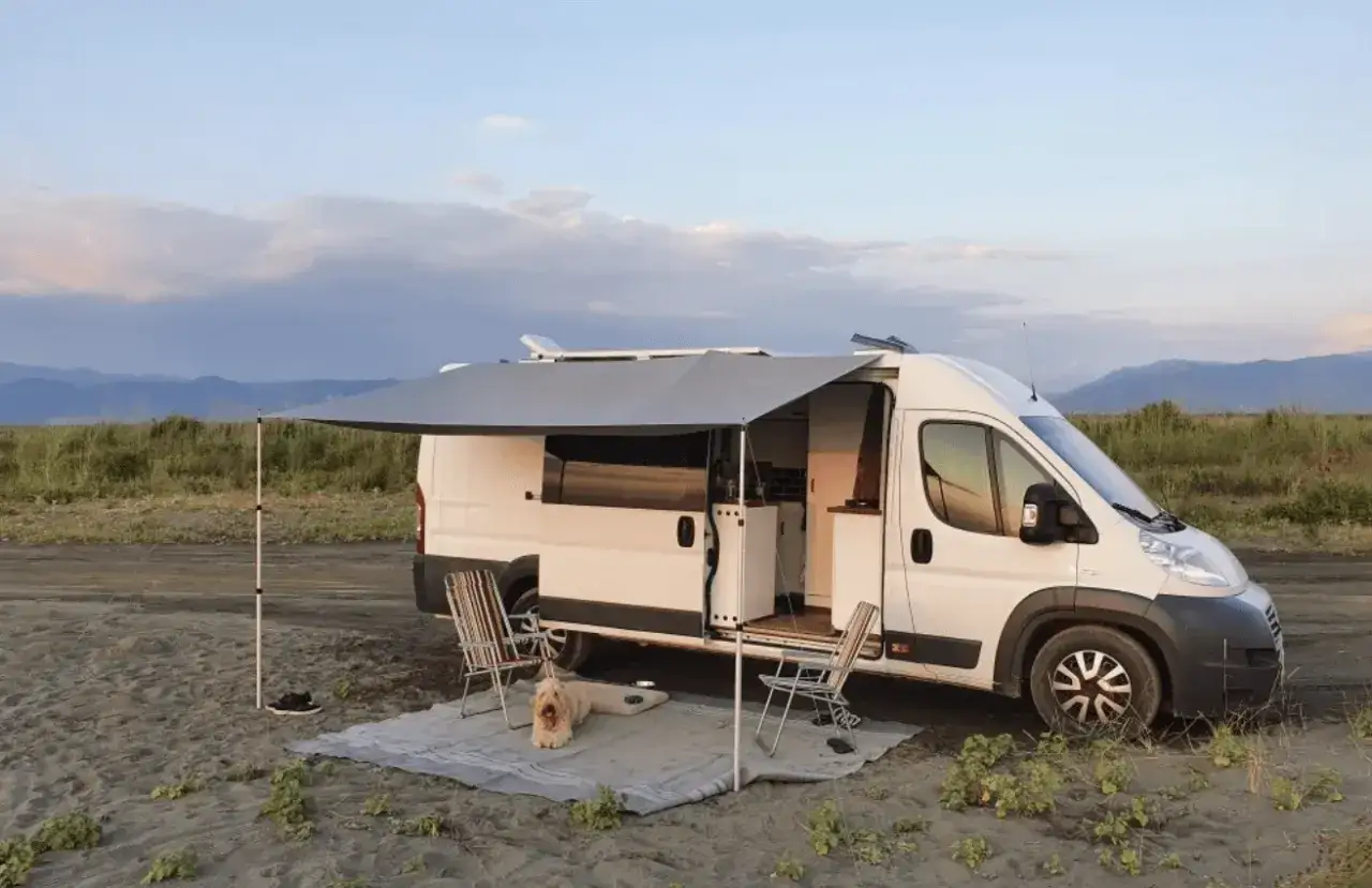Biały kamper 3 osobowy z rozłożonym markizą, dwoma krzesłami i psem na plaży.