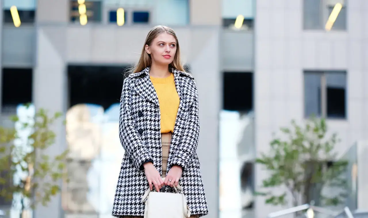 Kobieta w płaszczu w pepitkę i żółtym swetrze, z białą torebką. Czy pepitka jest jeszcze modna? Tak, wygląda świetnie!