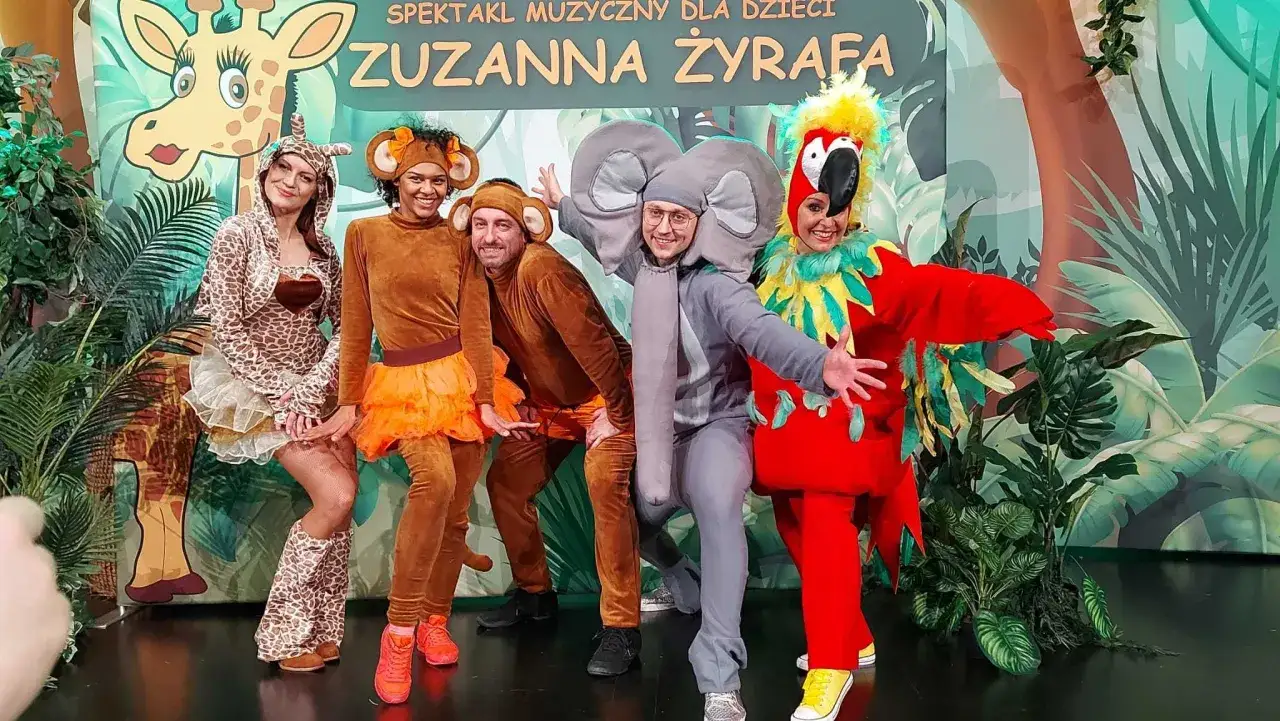 Aktorzy w kostiumach zwierząt, w tym słonia i papugi, pozują na tle scenografii z napisem 