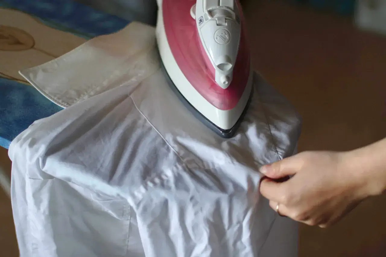 Dłoń układa białą koszulę na desce do prasowania, przygotowując ją do prasowania rękawów.