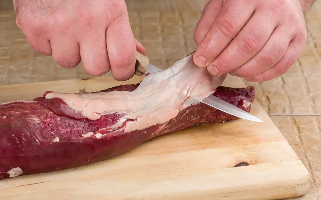 Ręce kroją błony i ścięgna z surowego mięsa na desce. Proste triki, jak pozbyć się błon i ścięgien z mięsa, ułatwią przygotowanie potrawy.